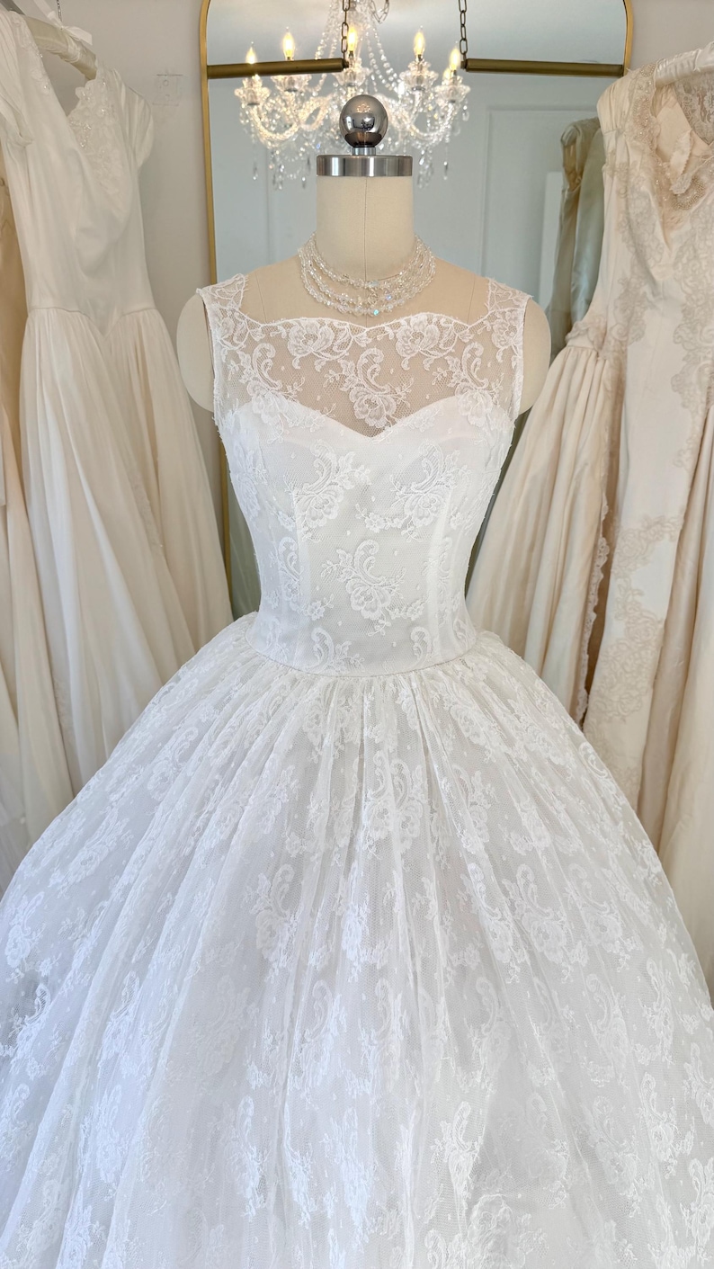 May include: A sleeveless white wedding dress featuring a sheer lace overlay with intricate floral patterns and delicate beadwork. The bodice has a sweetheart neckline under a high illusion lace yoke, transitioning into a full, voluminous skirt.