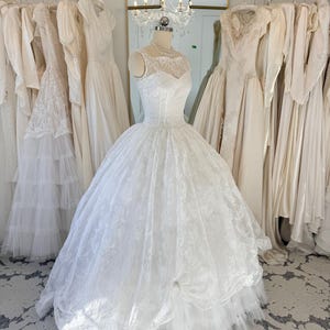 May include: A white sleeveless ball gown featuring a sweetheart neckline with a sheer lace overlay, a fitted bodice, and a voluminous floor-length skirt with intricate lace detailing and multiple layers of tulle fabric on a dress form.
