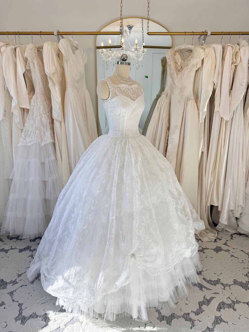 May include: A white sleeveless ball gown featuring a sweetheart neckline with a sheer lace overlay, a fitted bodice, and a voluminous floor-length skirt with intricate lace detailing and multiple layers of tulle fabric on a dress form.