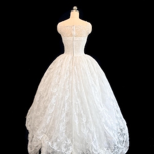 May include: A white floor-length gown featuring a fitted bodice with a sheer lace overlay and a full, voluminous skirt made of layered lace fabric. The back of the dress includes a central vertical zipper closure and a high, rounded neckline.