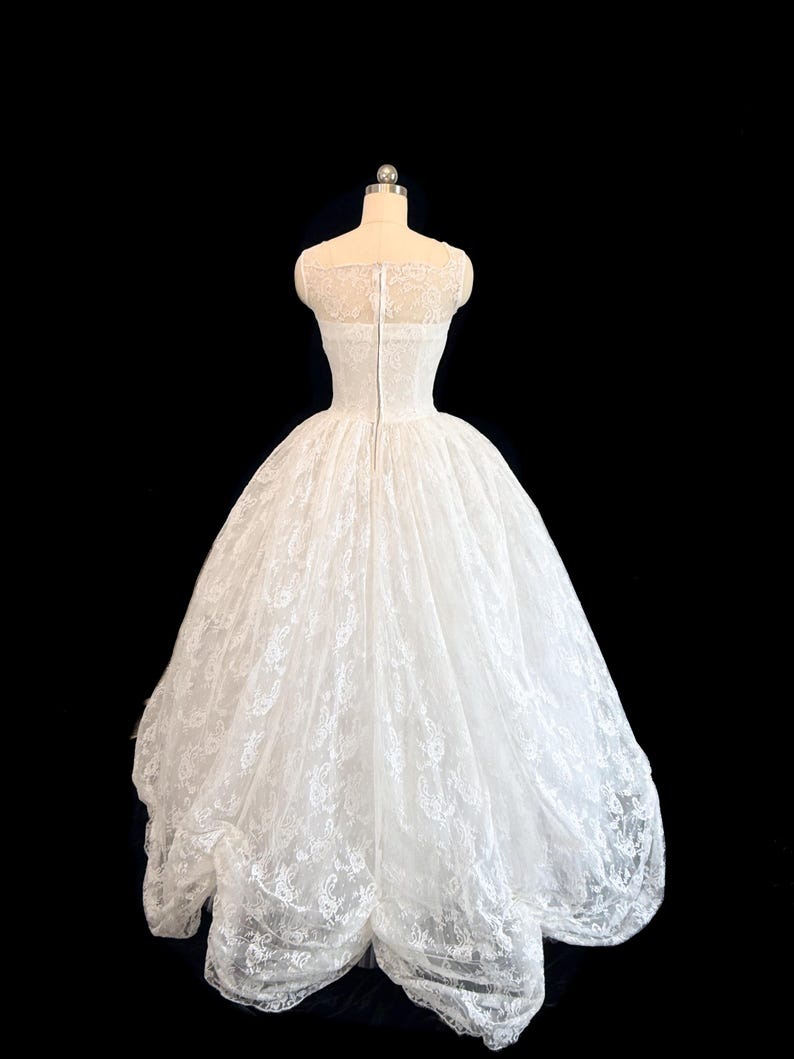 May include: A white floor-length gown featuring a fitted bodice with a sheer lace overlay and a full, voluminous skirt made of layered lace fabric. The back of the dress includes a central vertical zipper closure and a high, rounded neckline.