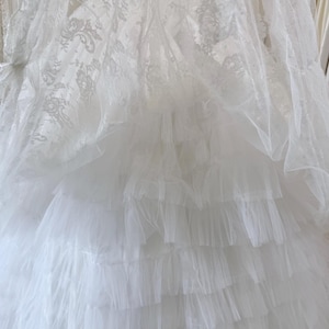 May include: A white dress featuring a top layer of sheer lace with an intricate floral pattern and a bottom section composed of multiple horizontal tiers of gathered tulle fabric, creating a voluminous and layered appearance throughout the garment.