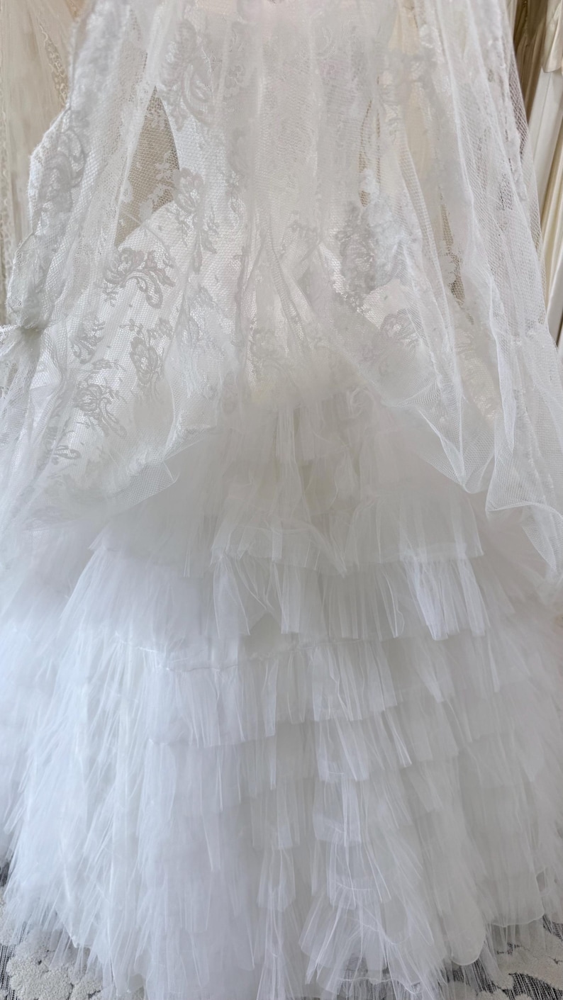 May include: A white dress featuring a top layer of sheer lace with an intricate floral pattern and a bottom section composed of multiple horizontal tiers of gathered tulle fabric, creating a voluminous and layered appearance throughout the garment.