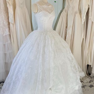 May include: A white sleeveless ball gown featuring a sheer lace illusion neckline and a fitted bodice. The voluminous skirt is crafted from layers of tulle and intricate floral lace overlay, creating a full silhouette that extends to the floor.