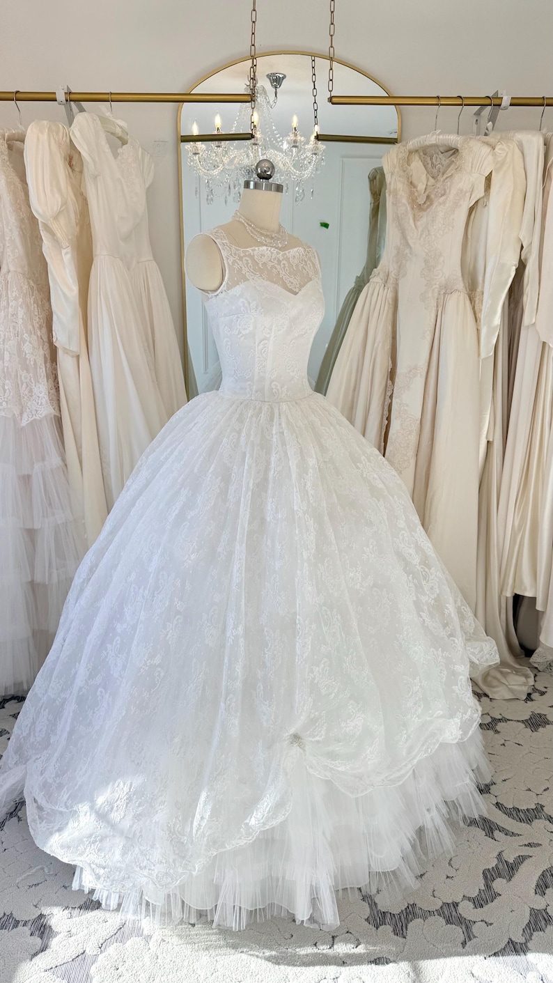 May include: A white sleeveless ball gown featuring a sheer lace illusion neckline and a fitted bodice. The voluminous skirt is crafted from layers of tulle and intricate floral lace overlay, creating a full silhouette that extends to the floor.