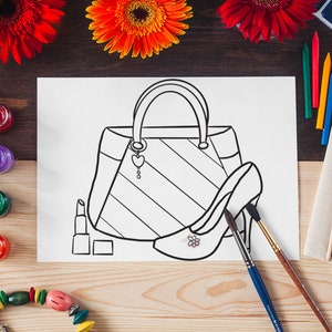 Puede incluir: Dibujo lineal en blanco y negro de un bolso, un zapato de tac&oacute;n y un l&aacute;piz labial sobre una hoja de papel blanca. El dibujo est&aacute; rodeado de materiales de arte, incluyendo pinceles, crayones y botes de pintura, con flores rojas y naranjas en el fondo.