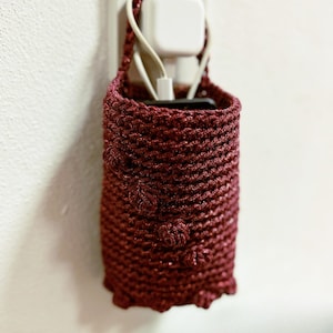 May include: A deep red crochet phone holder with a textured pattern and bobble detailing. Designed to hang from a wall socket, it holds a charging cable and a phone.