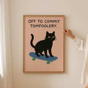 May include: A framed art print featuring a black cat on a blue skateboard. The cat has yellow eyes and is set against a pink background. The text at the top reads, "OFF TO COMMIT TOMFOOLERY."