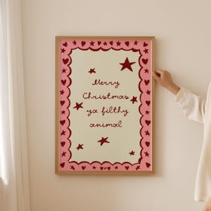 May include: A framed print with a light beige background and a pink border. The border is decorated with hearts and stars. The text "Merry Christmas ya filthy animal" is written in a cursive font in the center of the print. The frame is made of wood.