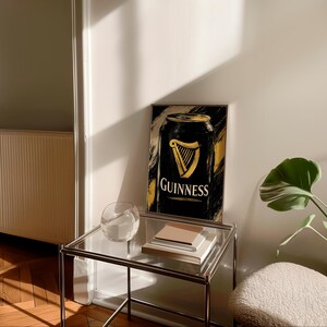 May include: A framed art print featuring a black Guinness can design with gold accents. The Guinness logo, a golden harp, is prominently displayed. The artwork is set against a textured, abstract background. The print is displayed on a glass-topped table.