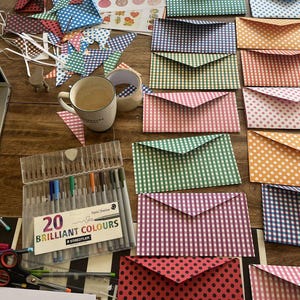 May include: A collection of colorful paper envelopes in various patterns, including polka dots and gingham. A set of 20 brilliant color pens and a coffee mug are also visible. The envelopes are in shades of red, pink, green, orange, and blue.