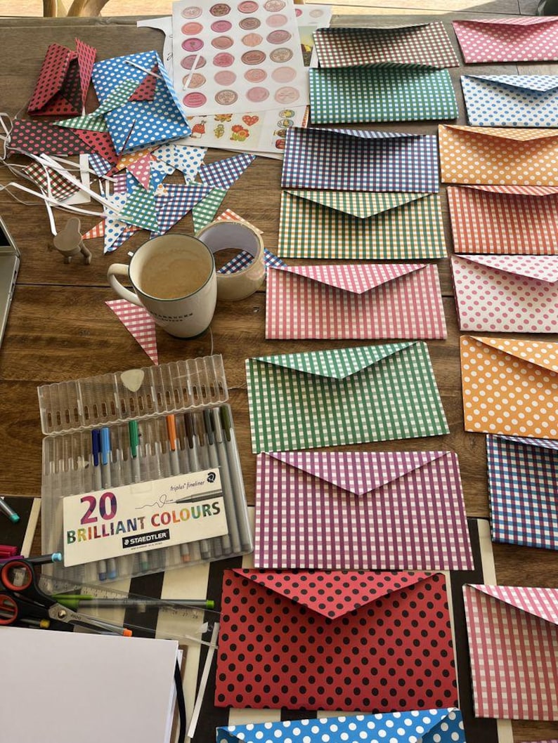 May include: A collection of colorful paper envelopes in various patterns, including polka dots and gingham. A set of 20 brilliant color pens and a coffee mug are also visible. The envelopes are in shades of red, pink, green, orange, and blue.