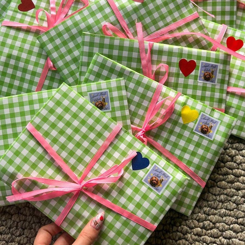 May include: A collection of gifts wrapped in green and white checkered paper, tied with pink ribbons. Each package is adorned with a heart-shaped sticker and a small stamp featuring a snail. The gifts are arranged on a textured surface.