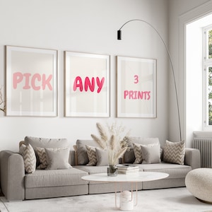 May include: Three framed prints with pink text on a beige background. The prints read "PICK", "ANY", and "3 PRINTS". The prints are hanging on a white wall above a gray couch with pillows.