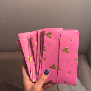 May include: A stack of pink envelopes with a gold heart pattern. The envelopes are held in a hand with a dark blue nail polish. The background is a neutral gray.