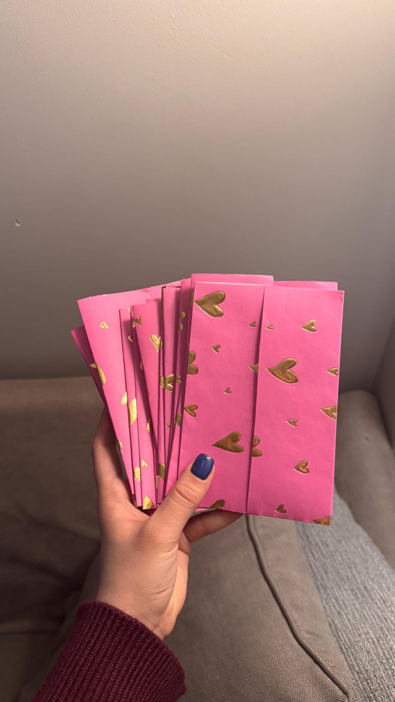 May include: A stack of pink envelopes with a gold heart pattern. The envelopes are held in a hand with a dark blue nail polish. The background is a neutral gray.