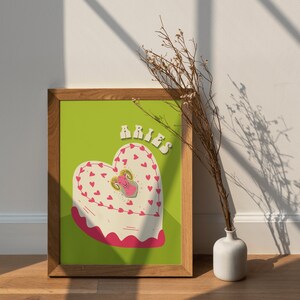 May include: A framed print with a green background and a white heart-shaped cake with pink frosting and red hearts. The cake has a cartoon ram on top and the word "Aries" in yellow letters at the top of the print.