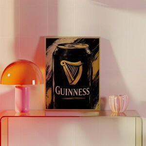 May include: A framed print featuring a stylized illustration of a Guinness can. The can is black with a gold harp logo and the word "Guinness" in white. The background has gold brushstrokes. The print is displayed on a glass shelf with a pink and orange lamp.