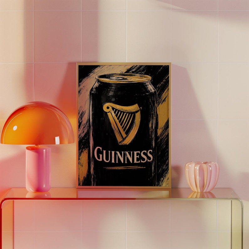 May include: A framed print featuring a stylized illustration of a Guinness can. The can is black with a gold harp logo and the word "Guinness" in white. The background has gold brushstrokes. The print is displayed on a glass shelf with a pink and orange lamp.