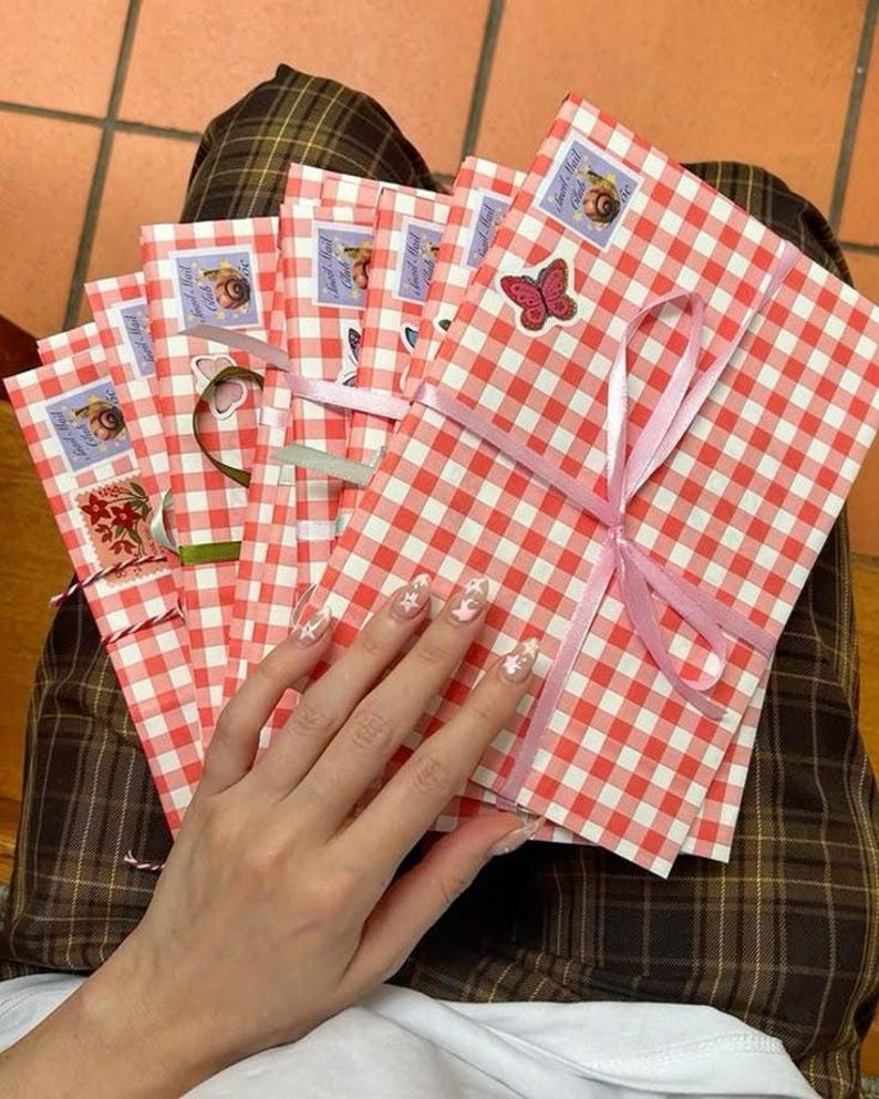 May include: A stack of red and white gingham patterned envelopes tied with pink ribbon. Each envelope is decorated with stickers, including a butterfly and a snail. The envelopes are held together.