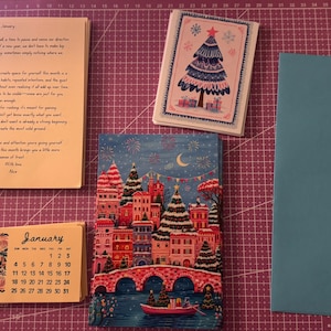 May include: A collection of stationery items, including a calendar, greeting cards, and an envelope, are displayed on a purple cutting mat. The calendar is labelled "January" and has a floral design. One card features a Christmas tree illustration.