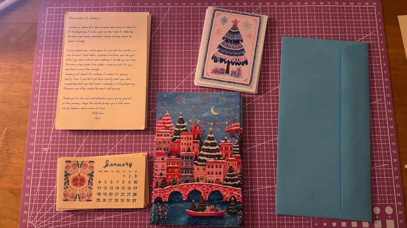 May include: A collection of stationery items, including a calendar, greeting cards, and an envelope, are displayed on a purple cutting mat. The calendar is labelled "January" and has a floral design. One card features a Christmas tree illustration.