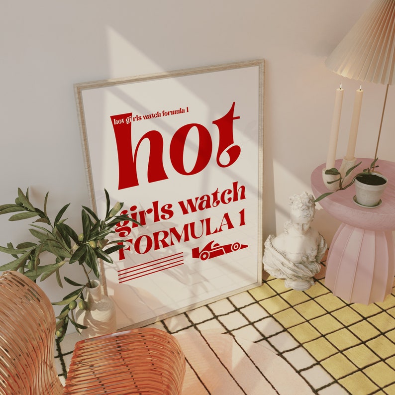 May include: A white framed poster with red text that reads "hot girls watch formula 1". The poster is hanging on a white wall with a light brown wooden floor.
