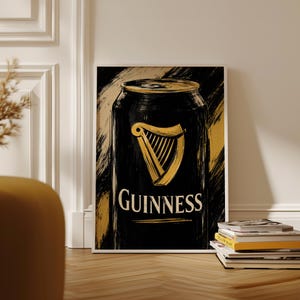 May include: A framed art print featuring a black Guinness can design with a gold harp logo and the Guinness text in white. The background has gold brushstrokes. The print is displayed on a wooden floor.