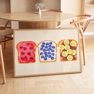 May include: A framed print of three slices of toast with different toppings. The first slice has red jam, the second has blueberries, and the third has banana slices and chocolate.