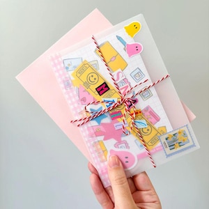 May include: A pink envelope with a vellum overlay tied with red and white twine. The overlay contains stickers and a patterned paper. The stickers include a yellow ticket with a smiley face, a pink ghost, and a blue stamp.