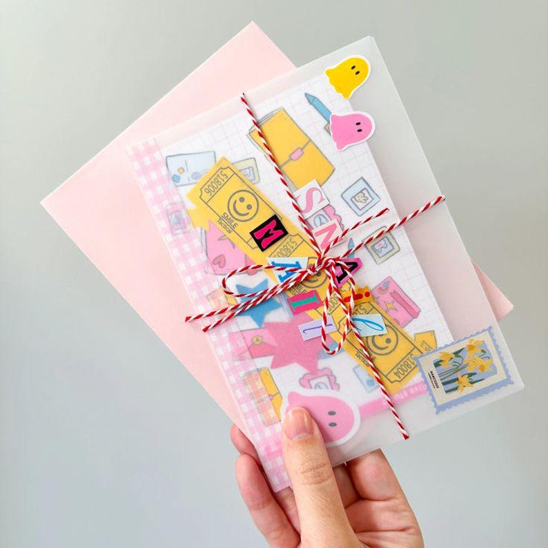 May include: A pink envelope with a vellum overlay tied with red and white twine. The overlay contains stickers and a patterned paper. The stickers include a yellow ticket with a smiley face, a pink ghost, and a blue stamp.