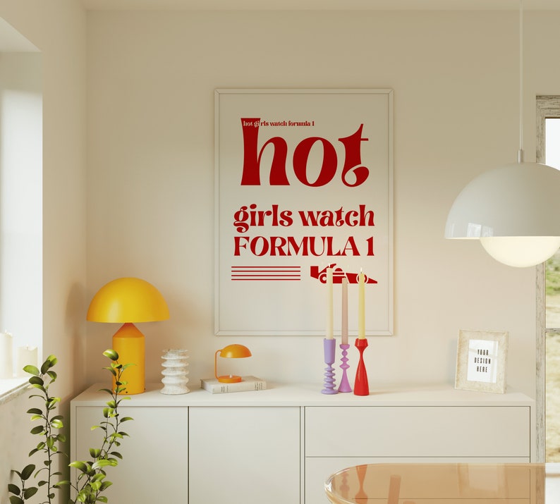 May include: A white framed poster with red text that reads "hot girls watch formula 1" with a red illustration of a race car. The poster is hanging on a white wall above a white dresser with a yellow lamp, a white decorative object, and a small book.