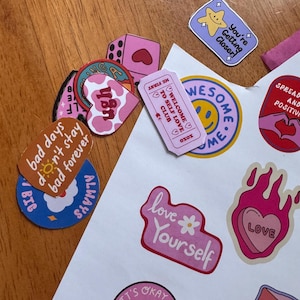 May include: A collection of colorful stickers with various designs and text. The stickers include phrases like "Love Yourself," "You're Getting Closer," and "It's Okay to Ask for Help." The color palette includes pink, blue, and yellow.
