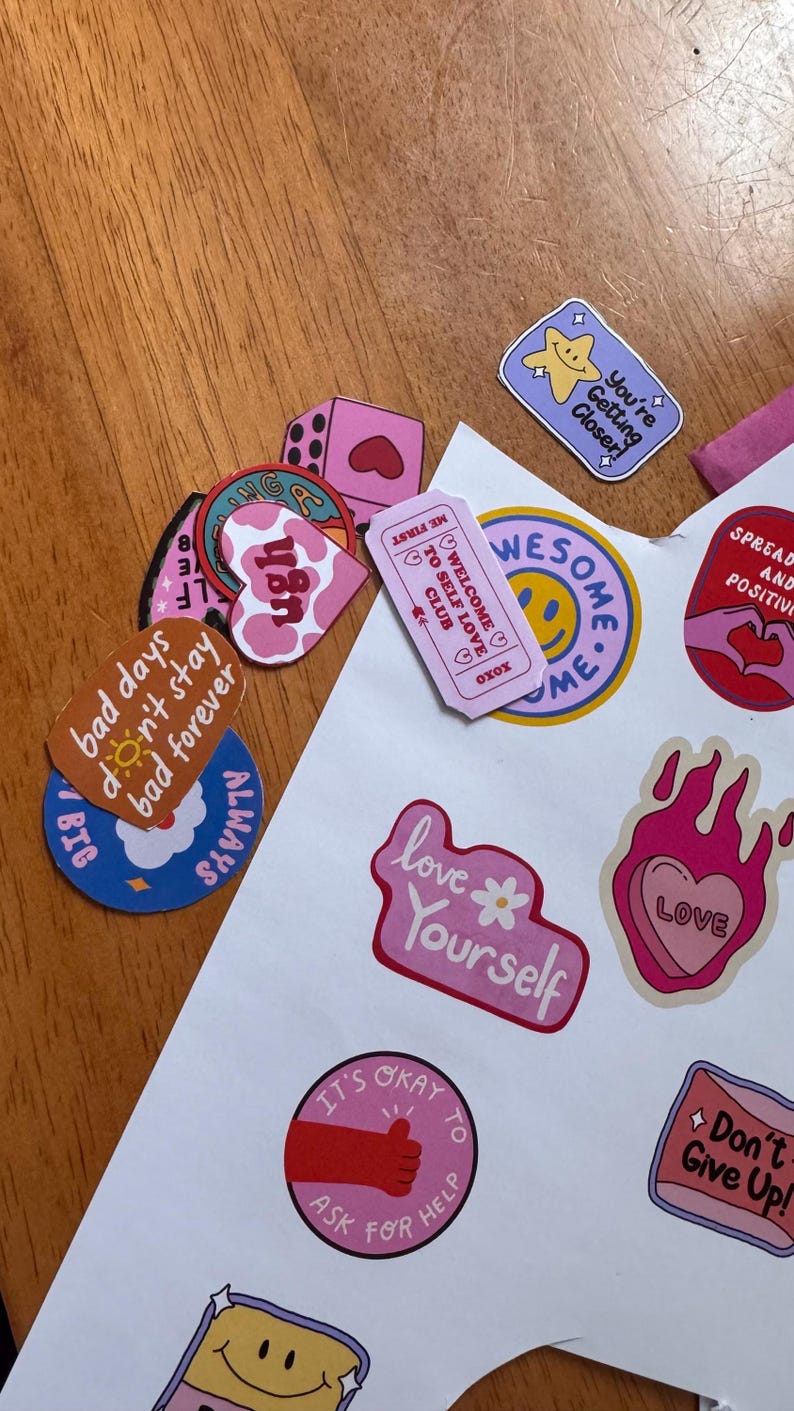 May include: A collection of colorful stickers with various designs and text. The stickers include phrases like "Love Yourself," "You're Getting Closer," and "It's Okay to Ask for Help." The color palette includes pink, blue, and yellow.