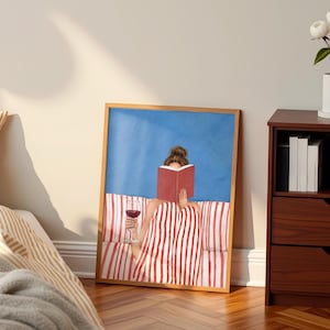 Peut inclure: Une affiche encadrée représente une personne lisant un livre et tenant un verre de vin rouge. L'œuvre d'art présente un fond bleu et un cadre en bois. La personne est assise sur une surface rayée. Un vase blanc avec des fleurs est posé sur une commode marron foncé.
