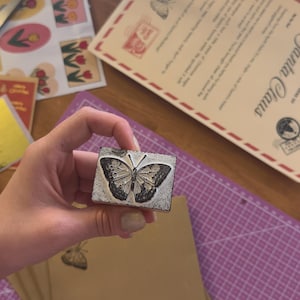 May include: A rectangular wooden stamp with a detailed butterfly design. The stamp is held over a stack of brown paper and a purple cutting mat. Other stationery items are visible in the background.