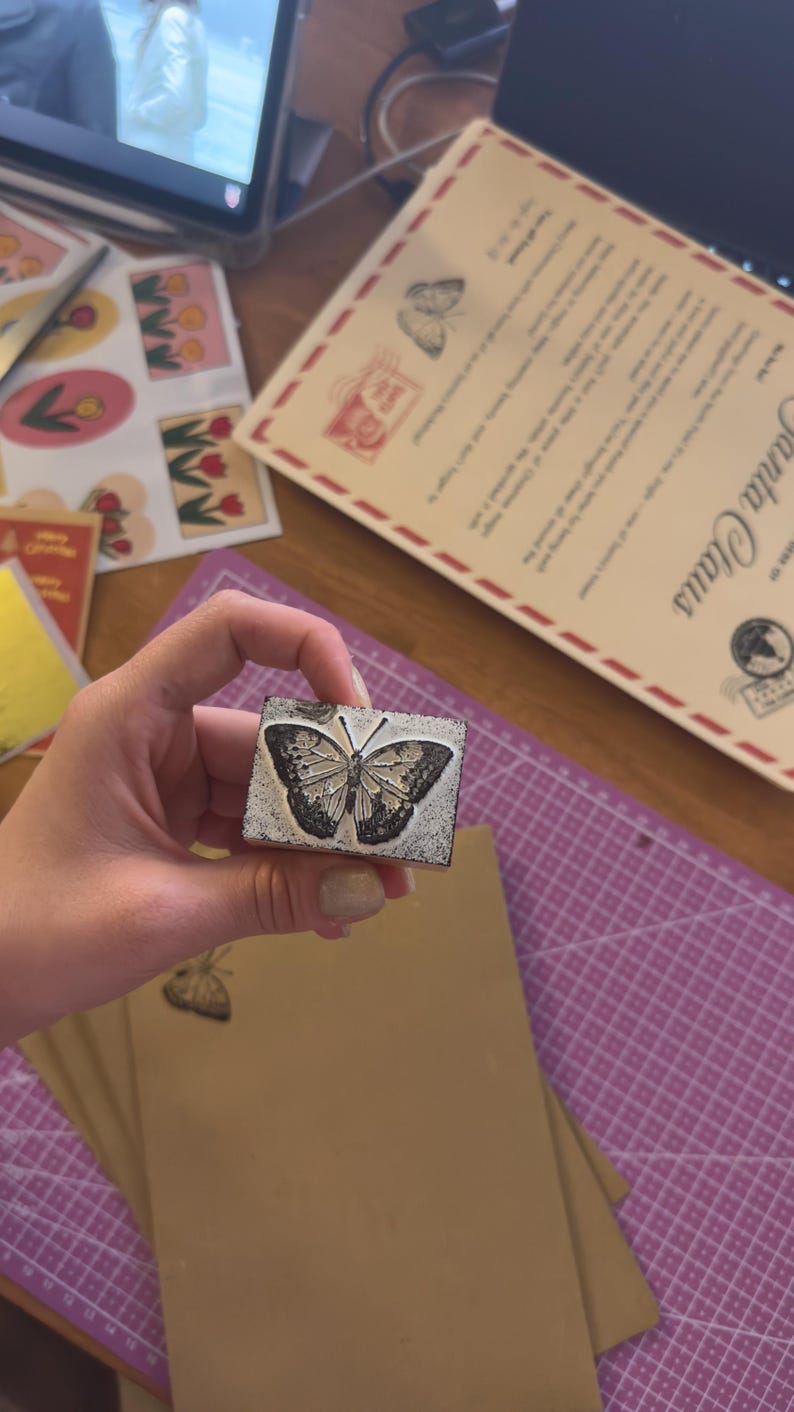 May include: A rectangular wooden stamp with a detailed butterfly design. The stamp is held over a stack of brown paper and a purple cutting mat. Other stationery items are visible in the background.