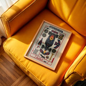 May include: A framed print featuring a Red Bull Racing 2018 Formula 1 car design. The artwork, with a light wood frame, is displayed on a yellow upholstered armchair. The print includes the Red Bull logo and car components.