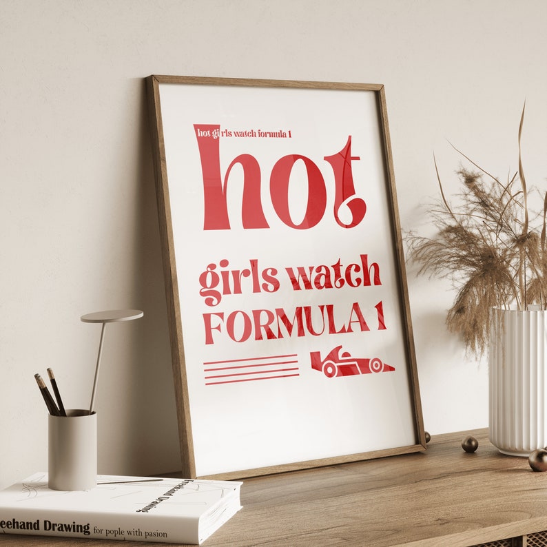 May include: A white framed print with red text that reads "hot girls watch formula 1". The text "hot" is in a large, bold font. A small red race car graphic is below the text.