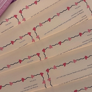 May include: Several cream-colored cards with a decorative border of pink and red hearts connected by a black line. The cards have text in a script font. A pink paper cutter is visible in the upper left corner.
