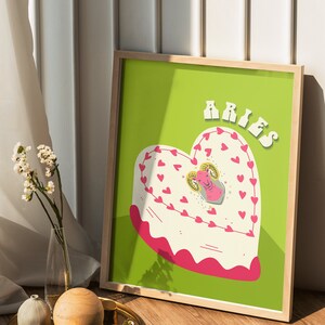May include: A framed print with a green background featuring a heart-shaped cake with pink frosting and a white ram illustration in the center. The text "ARIES" is written in pink letters at the top of the print.