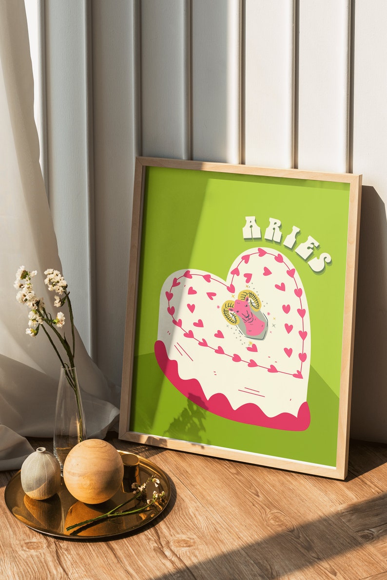 May include: A framed print with a green background featuring a heart-shaped cake with pink frosting and a white ram illustration in the center. The text "ARIES" is written in pink letters at the top of the print.