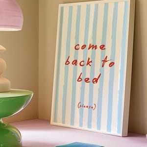 Puede incluir: Una impresión enmarcada blanca con rayas azules y blancas. La impresión tiene el texto "come back to bed (please)" en rojo.