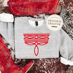 May include: A heather gray sweatshirt with a red embroidered design, displayed with red cowboy boots and a leopard print scarf. The sweatshirt features a unique, symmetrical design. The image also includes the text "ORIGINAL CREATOR".