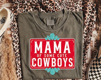 May include: Olive green t-shirt with a red rectangular graphic that reads "MAMA OF SOME CUTE COWBOYS" in white text. The graphic is decorated with turquoise teardrop and petal shapes. The shirt is laid on leopard print pants and a pair of Vans shoes.