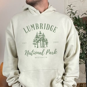 May include: A light beige hoodie with a green graphic print of a forest and the text "Lumbridge National Park Est. 2001 Mistahalin".
