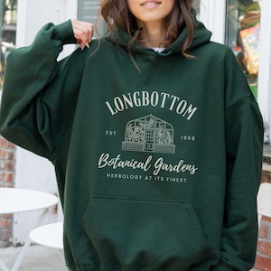 May include: Dark green hooded sweatshirt with a front pocket. The sweatshirt features the text "LONGBOTTOM BOTANICAL GARDENS" in white, along with a greenhouse illustration. The text also includes "EST. 1990" and "HERBOLOGY AT ITS FINEST".