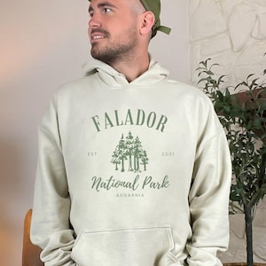 May include: A light beige hooded sweatshirt with a green graphic print that says "Falador National Park Asgaarnia" with the text "Est. 2001" above the graphic and "Est. 2001" below the graphic.