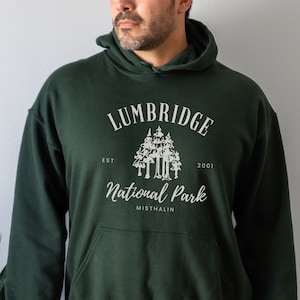 May include: A dark green hooded sweatshirt with a white graphic design that says "Lumbridge National Park Est. 2001 Misthalin". The graphic features a stylized image of a group of trees.