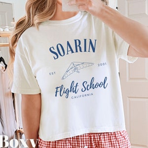 May include: White boxy t-shirt with a blue graphic of a hang glider and the text "Soarin' Flight School Est. 2001 California".